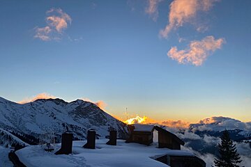 Snow-covered rooftops and chimneys overlook majestic mountains glowing under a golden sunset.