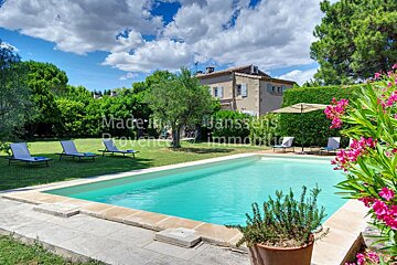 A large swimming pool is surrounded by chairs and umbrellas in front of a house that says janssens immobilier
