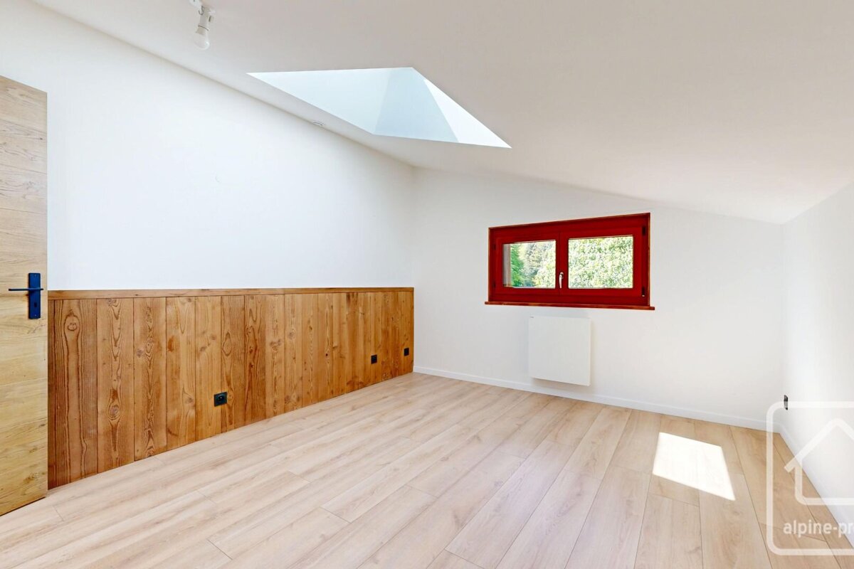 Bright, empty room with light wood floors, white walls, lower wood paneling, a red-framed window, and a skylight.