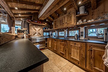 A cozy chalet kitchen with rich wooden beams, cabinets, and black countertops, featuring a stainless steel stove and scenic windows.