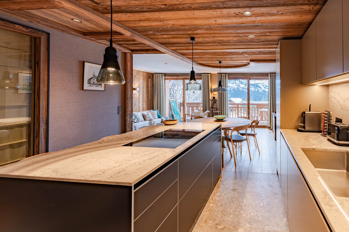 Modern chalet kitchen with a marble island and wooden ceiling, opening to a dining area with mountain views.