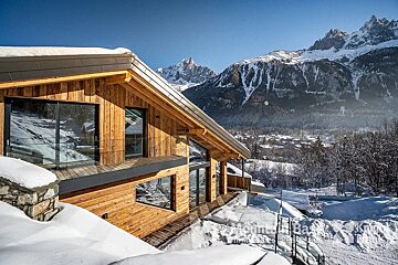 A wooden house with mountains in the background is covered in snow