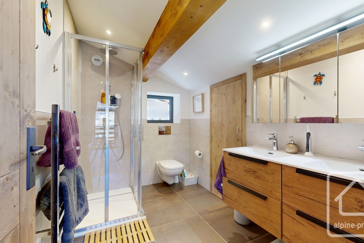 A modern rustic bathroom featuring a glass shower, wall-mounted toilet, and double wooden vanity. Light tiles and a wooden ceiling beam complete the look.