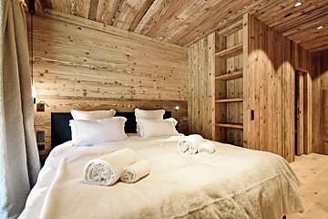 Cozy rustic bedroom with wooden paneled walls and ceiling, featuring a bed made with white linens and rolled towels.