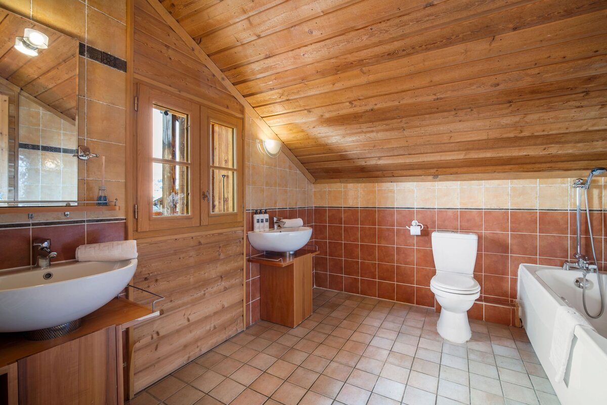 Rustic chalet bathroom with a slanted wooden ceiling, two modern sinks, a toilet, a bathtub, and warm tiled walls.