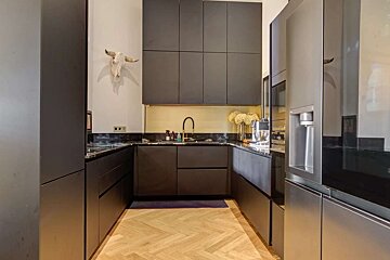 A modern U-shaped kitchen features sleek dark cabinets, black marble countertops, and light herringbone flooring. A bull skull and gold accents add character.