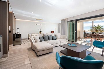 A bright, modern living room with a cream sectional sofa, leading to a balcony with outdoor seating and a stunning ocean view.