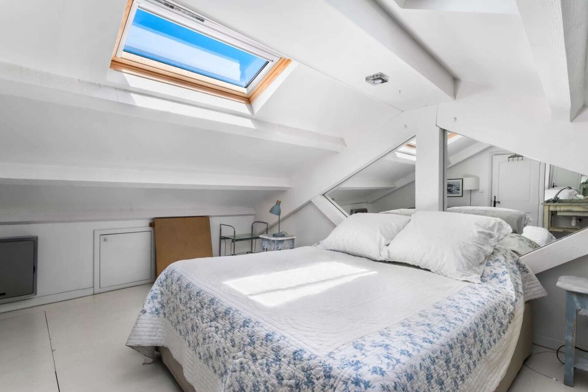 A bright, white attic bedroom featuring a skylight, a bed with a blue and white patterned duvet, and a mirrored wall reflecting the space.