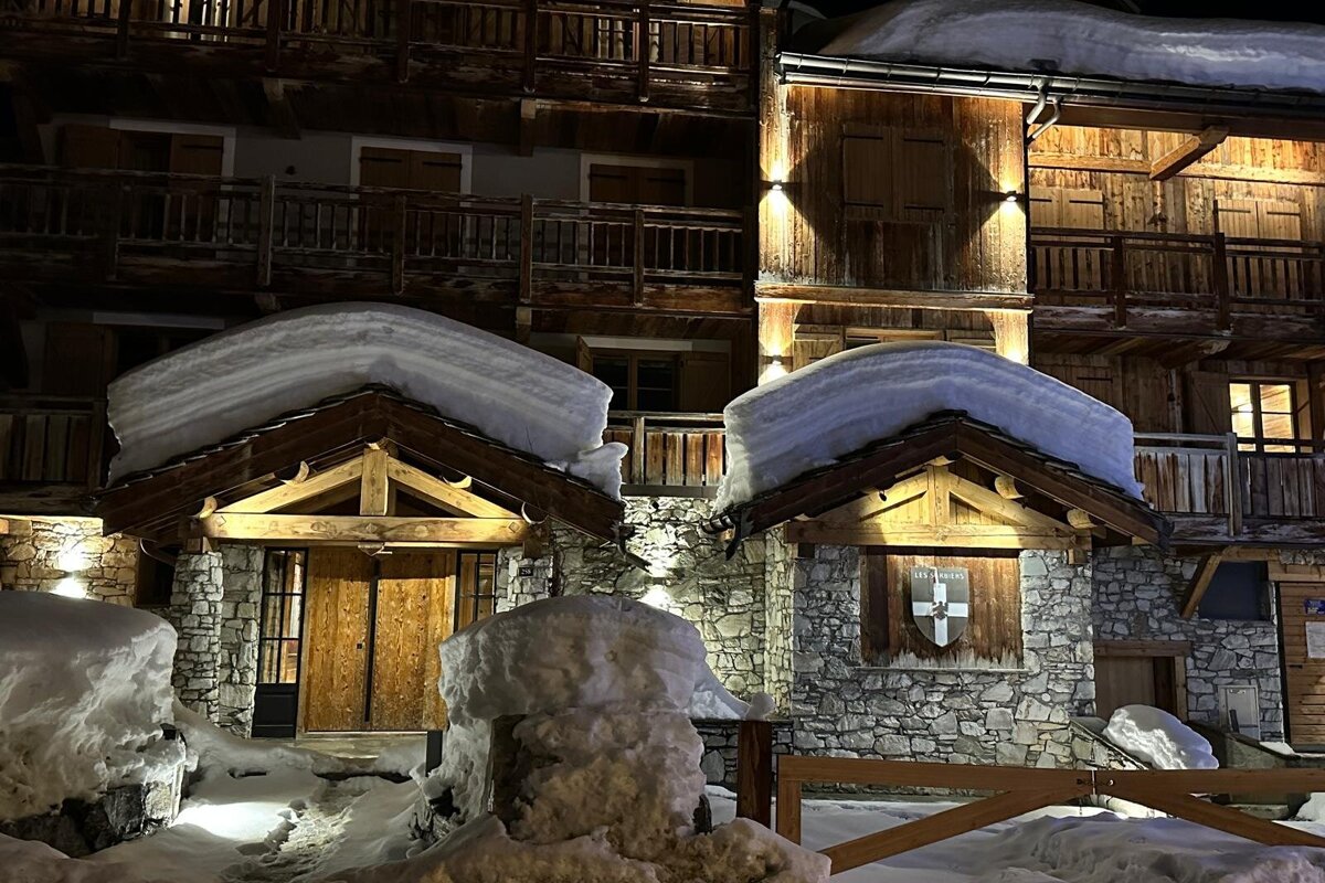 A charming, snow-covered wooden and stone building illuminated at night, with thick snow cascading from its roofs and piled around its entrances.