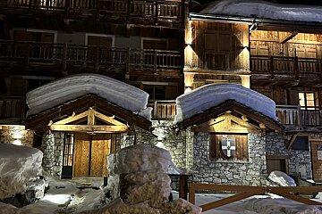 A charming, snow-covered wooden and stone building illuminated at night, with thick snow cascading from its roofs and piled around its entrances.