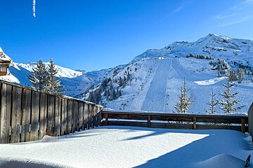 A snow-covered balcony overlooks sunny ski slopes with lifts & pine trees in majestic mountains under a bright blue sky.