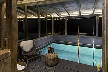 Cozy, rustic indoor pool area with exposed wooden beams. Champagne, glasses, and a fur-covered chair invite relaxation next to the softly lit water.