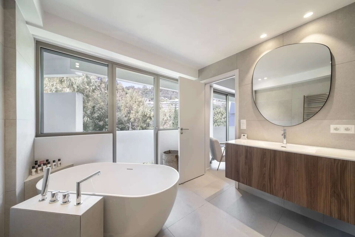 A modern bathroom features a freestanding tub, large windows with a view of lush greenery, and a sleek wooden vanity with a round mirror.