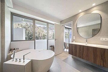 A modern bathroom features a freestanding tub, large windows with a view of lush greenery, and a sleek wooden vanity with a round mirror.