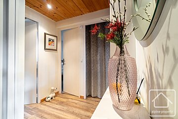 A cozy hallway with a wooden ceiling, light floor, white walls, and multiple doors. Decor includes a pink textured vase with flowers, a round mirror, and a small dog figure.