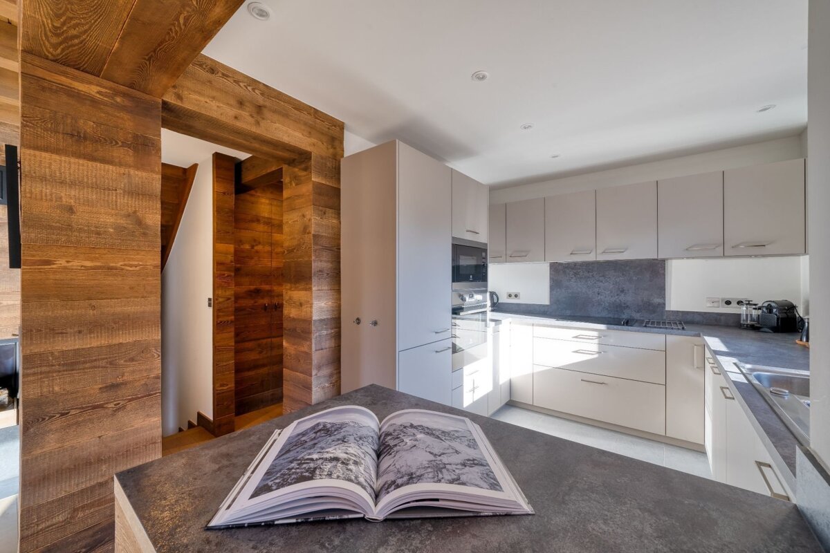 A modern kitchen with light cabinets and dark counters, featuring rustic wooden walls and a visible staircase. An open book rests on the foreground counter.