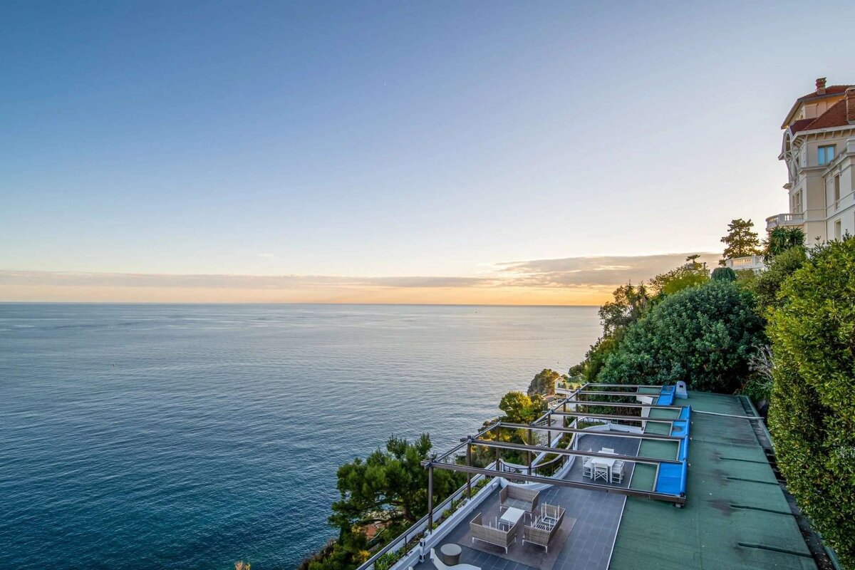 A grand cliffside mansion and terrace overlook a tranquil sea under a clear sky, with soft sunset hues on the horizon.