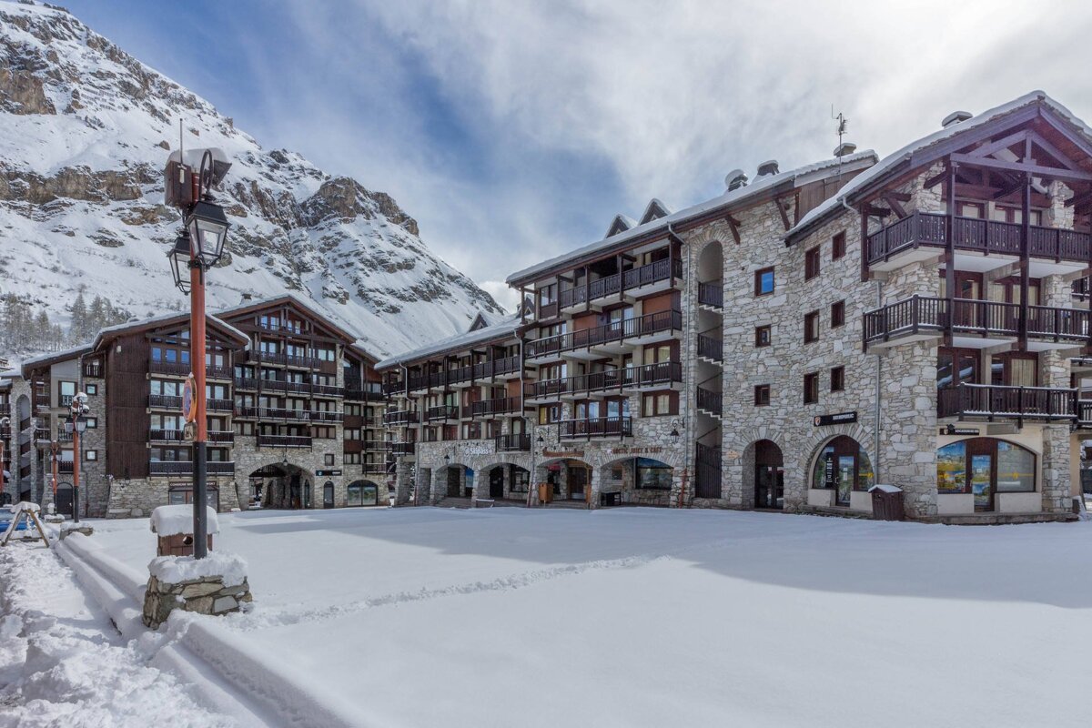 A snowy alpine village square surrounded by multi-story stone and timber buildings, with majestic snow-capped mountains in the background.