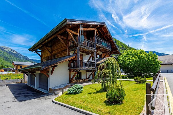 A large, multi-story wooden chalet with balconies and attached garages stands on a green lawn, surrounded by lush mountains under a bright blue sky.