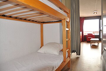 A room with wooden bunk beds on the left, separated by a curtain from a living area featuring a red armchair, small table, and a large window.