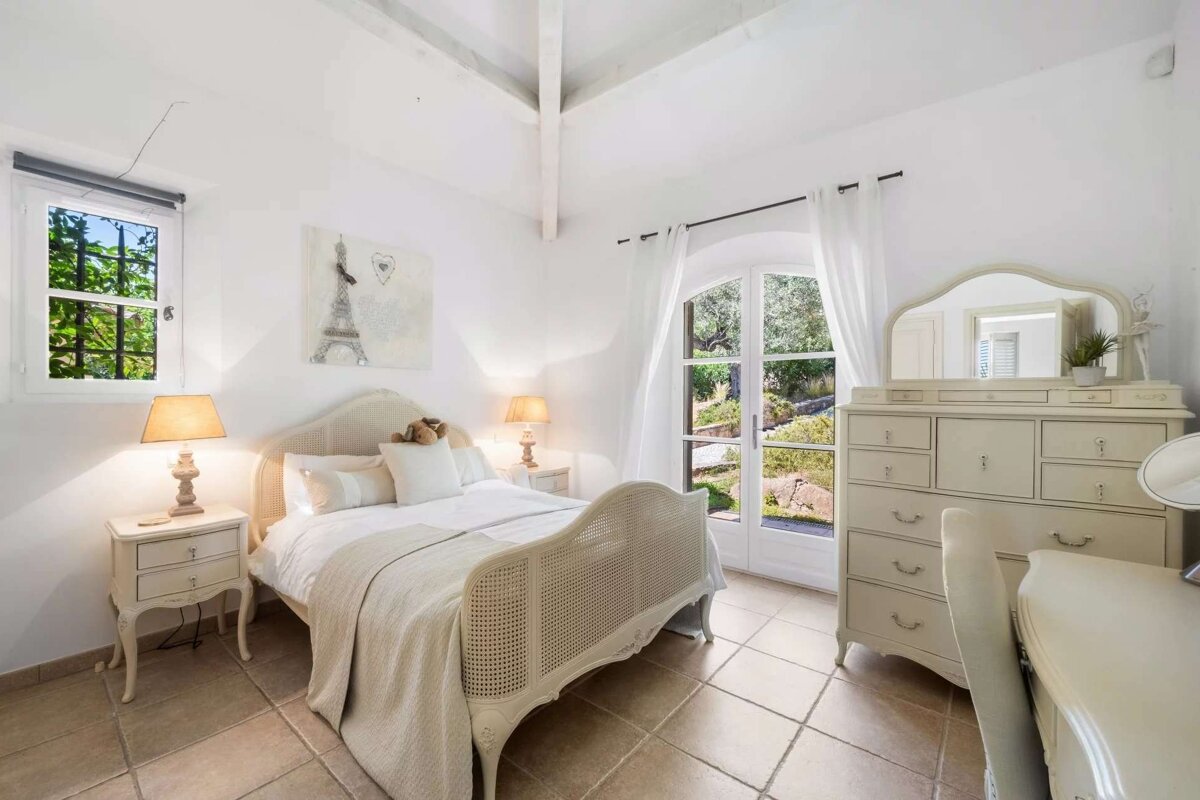 A bright, elegant bedroom features a white bed, nightstands with lamps, a dresser, and a window and glass door opening to a lush garden. White walls and tiled floors complete the chic look.