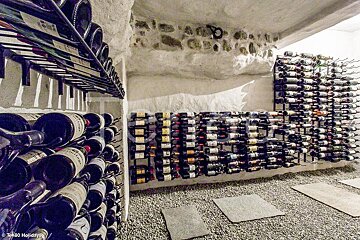 A rustic wine cellar with white stone walls, gravel flooring, and racks filled with wine bottles.