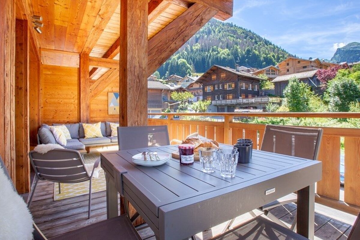 A cozy wooden chalet balcony with a dining table and lounge area, overlooking a scenic alpine mountain village.