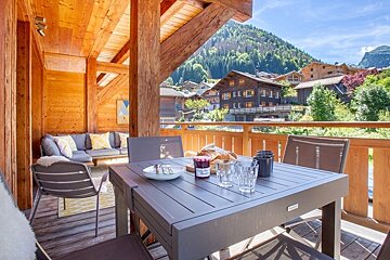 A cozy wooden chalet balcony with a dining table and lounge area, overlooking a scenic alpine mountain village.