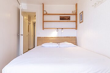 A clean bedroom with a bed, wicker headboard, and wooden shelves on a white wall. A doorway on the left leads to a hallway with closets.