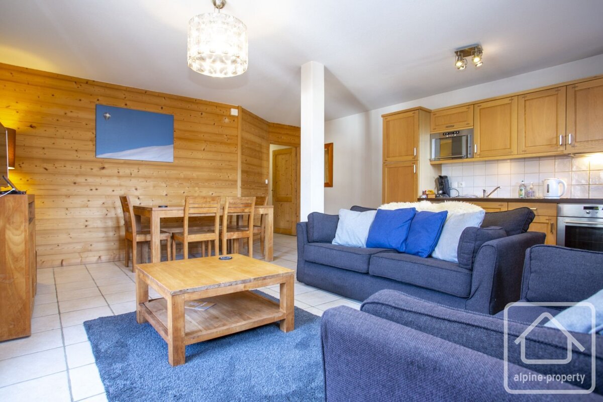 Rustic yet modern open-plan living space with wood walls, blue sofas and rug, a dining table, and a functional kitchen area.