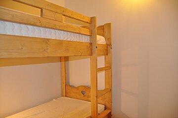 A wooden bunk bed with white mattresses stands against a pale wall. The lower bunk features a heart-shaped cut-out on its headboard.
