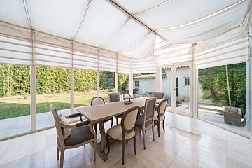 Elegant sunroom featuring a dining set for eight, marble floors, and panoramic windows with white blinds. A lush garden is visible outside under a fabric-draped ceiling.