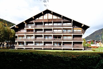 A large, multi-story chalet-style building with brown wooden balconies and a pointed roof, set against mountains. A green hedge is in the foreground.