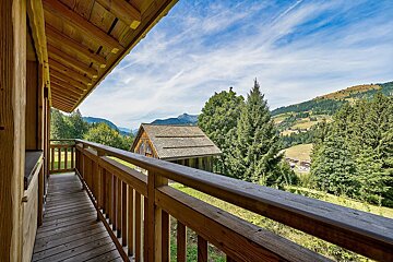 Scenic mountain view from a wooden balcony, overlooking green hills, pine trees, and a rustic barn under a bright blue sky.