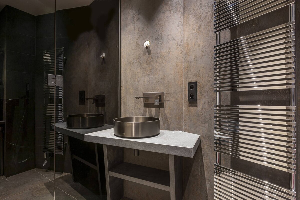 Modern industrial bathroom with a metallic vessel sink on a grey vanity, concrete-style walls, and a large chrome towel dryer.