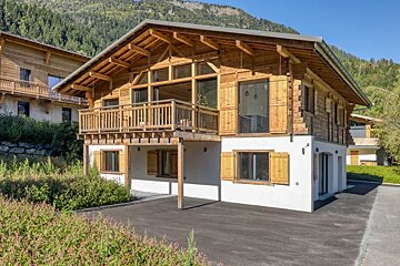 A two-story alpine chalet with a wooden upper level and balcony, a white ground floor, and a mountain backdrop.