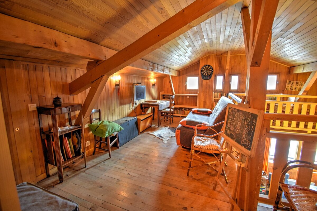 A cozy, rustic all-wood attic room featuring exposed beams, a sofa, TV, dartboard, table, chairs, and a chalkboard, illuminated by warm lighting.