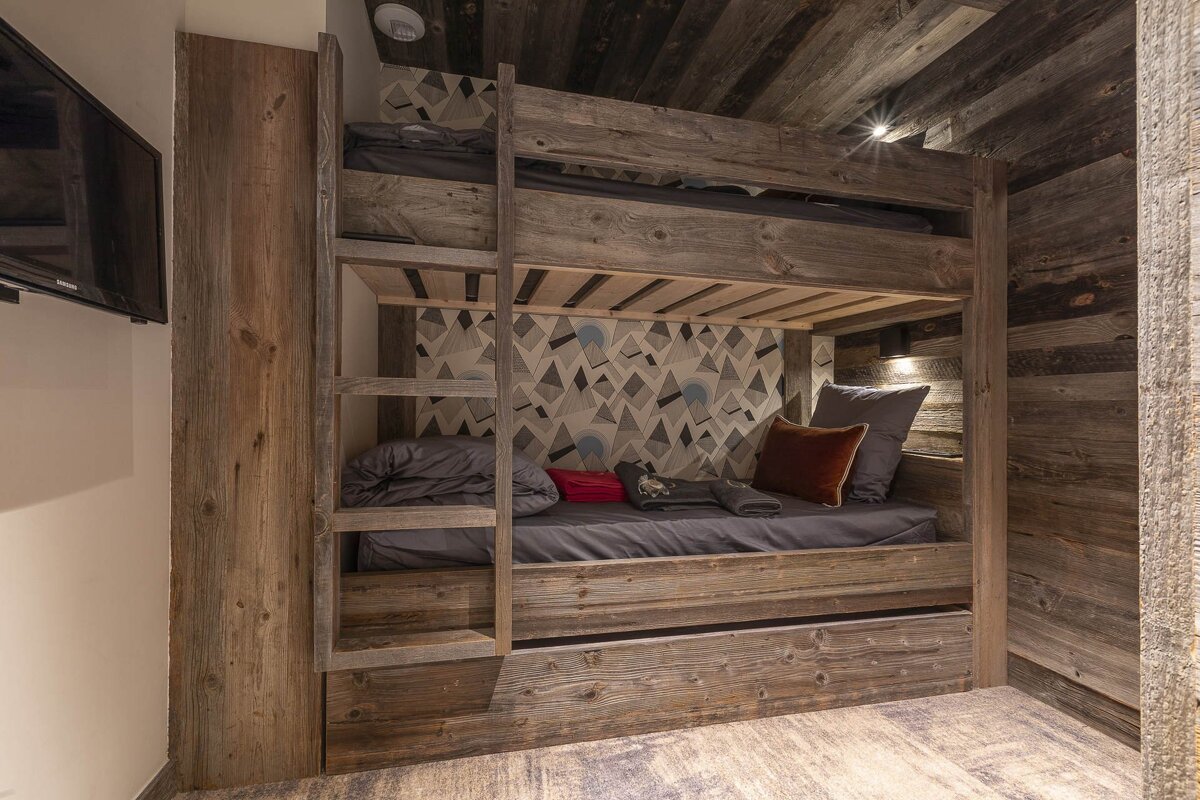 Rustic bunk bed room with weathered wood walls & ceiling, geometric wallpaper, cozy bedding, and a wall-mounted TV. Modern mountain chalet style.