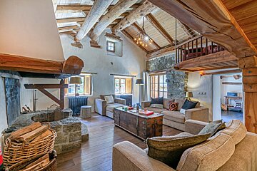Rustic living room with high log-beam ceilings, stone fireplace, beige sofas, a wooden trunk coffee table, and an overhead loft area.