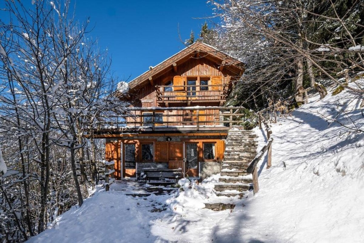 A rustic wooden chalet with balconies and stone steps is nestled in a snow-covered mountain, surrounded by trees under a clear blue sky.