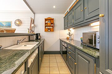 A narrow galley kitchen featuring dark gray cabinets, green marble countertops, white subway tiles, and a tiled floor. Appliances, a wooden shelf, and decorative skis are visible.