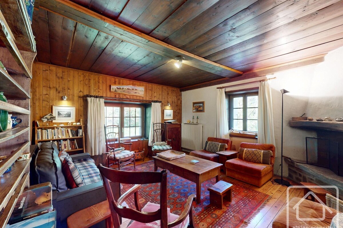 A cozy, rustic living room with wood-paneled walls and ceiling, a stone fireplace, traditional furniture, and bookshelves by sunlit windows.