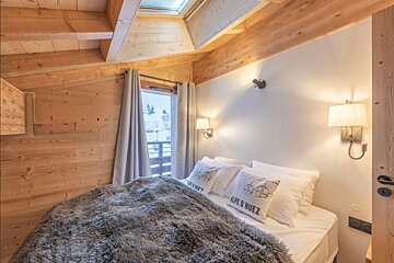 Cozy chalet bedroom with wooden paneled walls, a plush grey throw blanket, warm wall lamps, and a view of a snowy landscape.