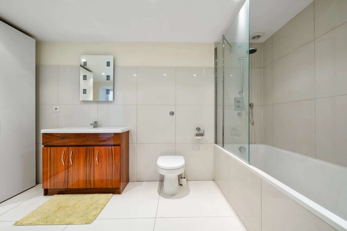 A modern bathroom with white tiles features a dark wood vanity with a sink, a toilet, and a bathtub with a glass shower screen. A yellow rug sits on the floor.