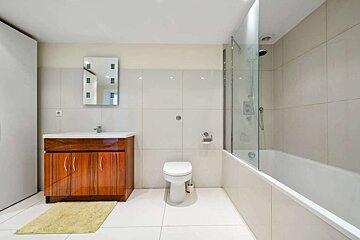 A modern bathroom with white tiles features a dark wood vanity with a sink, a toilet, and a bathtub with a glass shower screen. A yellow rug sits on the floor.