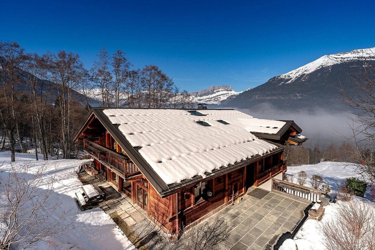 A rustic wooden chalet with a snow-covered roof sits amidst a winter wonderland. Snowy mountains rise in the background under a clear, bright blue sky.