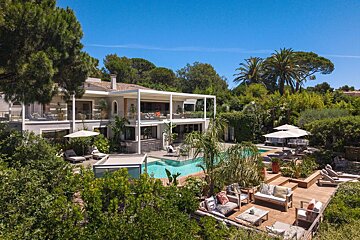 Luxurious modern villa featuring a swimming pool, outdoor seating decks, and lush tropical greenery under a clear blue sky.
