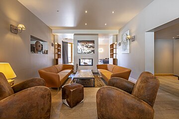 Cozy modern lounge with brown leather armchairs, orange sofas facing a coffee table, and a central fireplace.