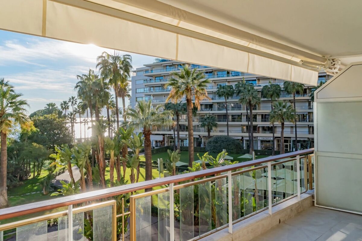 Sunny balcony view overlooking lush gardens, palm trees, and a modern resort building.