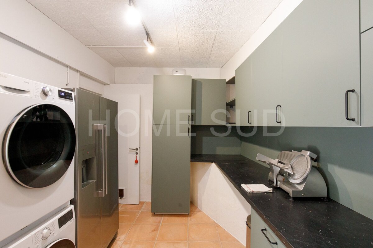 A functional utility kitchen featuring stacked laundry appliances, a fridge, sage green cabinets, dark counters with a meat slicer, and terracotta floors.